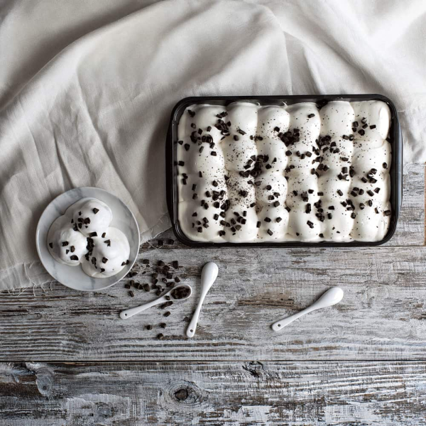 Profiteroles bianchi in vaschetta, ricoperti da crema chiara e decorati con scaglie di cioccolato, presentati in un’elegante ambientazione da dessert. Venduto da Orobica Food.