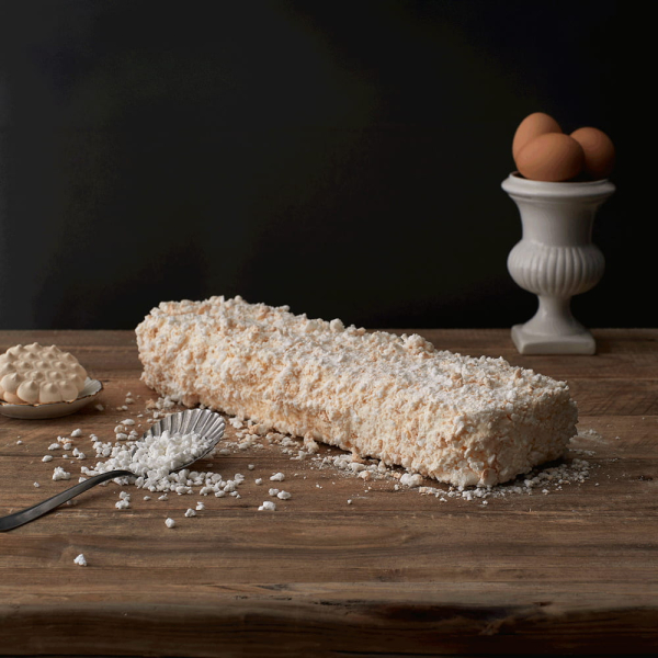 Rotolo di torta meringata ricoperto di briciole e zucchero a velo, con ingredienti e dettagli rustici su piano in legno, ideale per dessert artigianali. Venduto da Orobica Food.